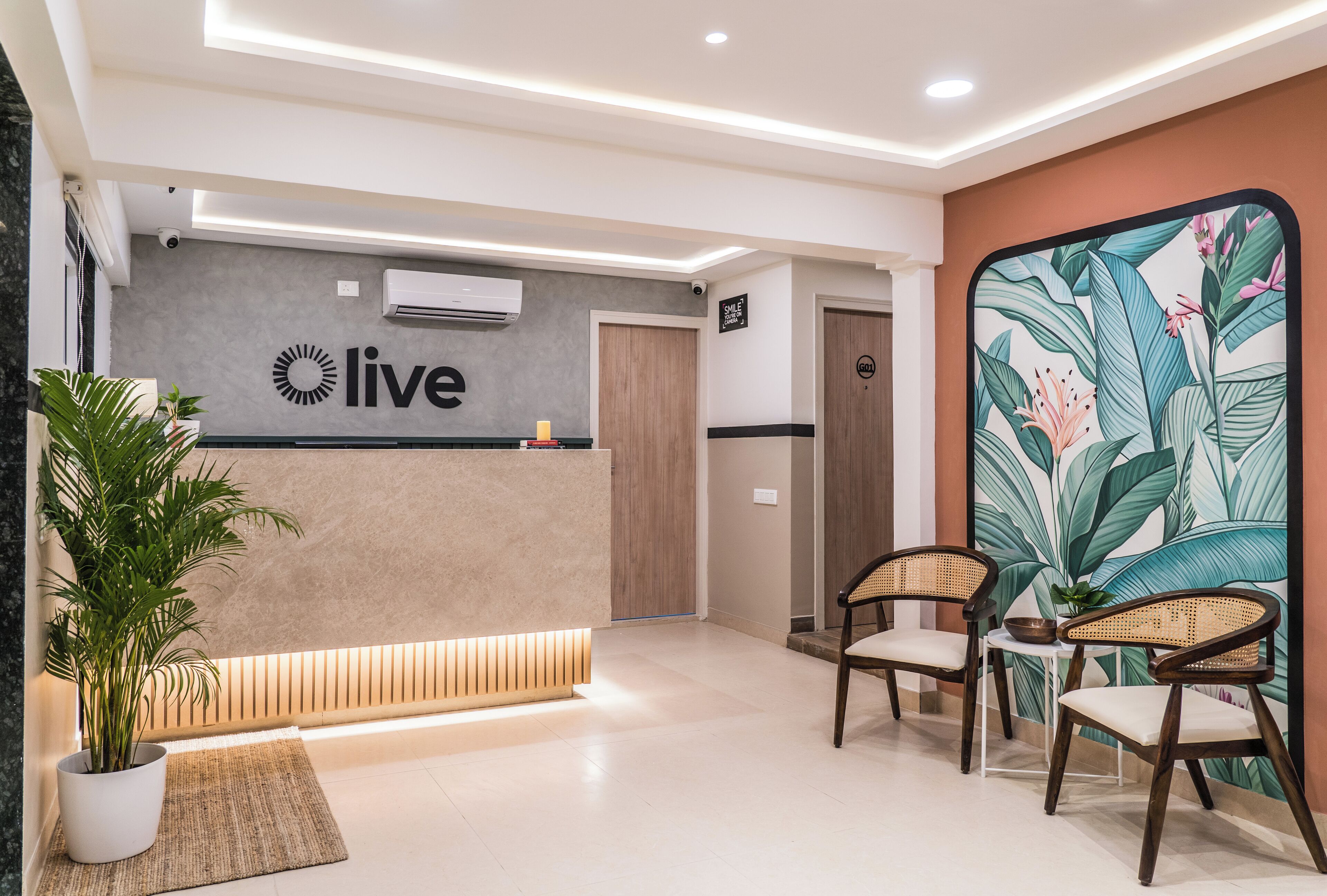 Un espace d’accueil avec un panneau indiquant « accueil », un comptoir de réception, deux fauteuils en osier, une grande plante tropicale et un mur orné d’un motif végétal.
