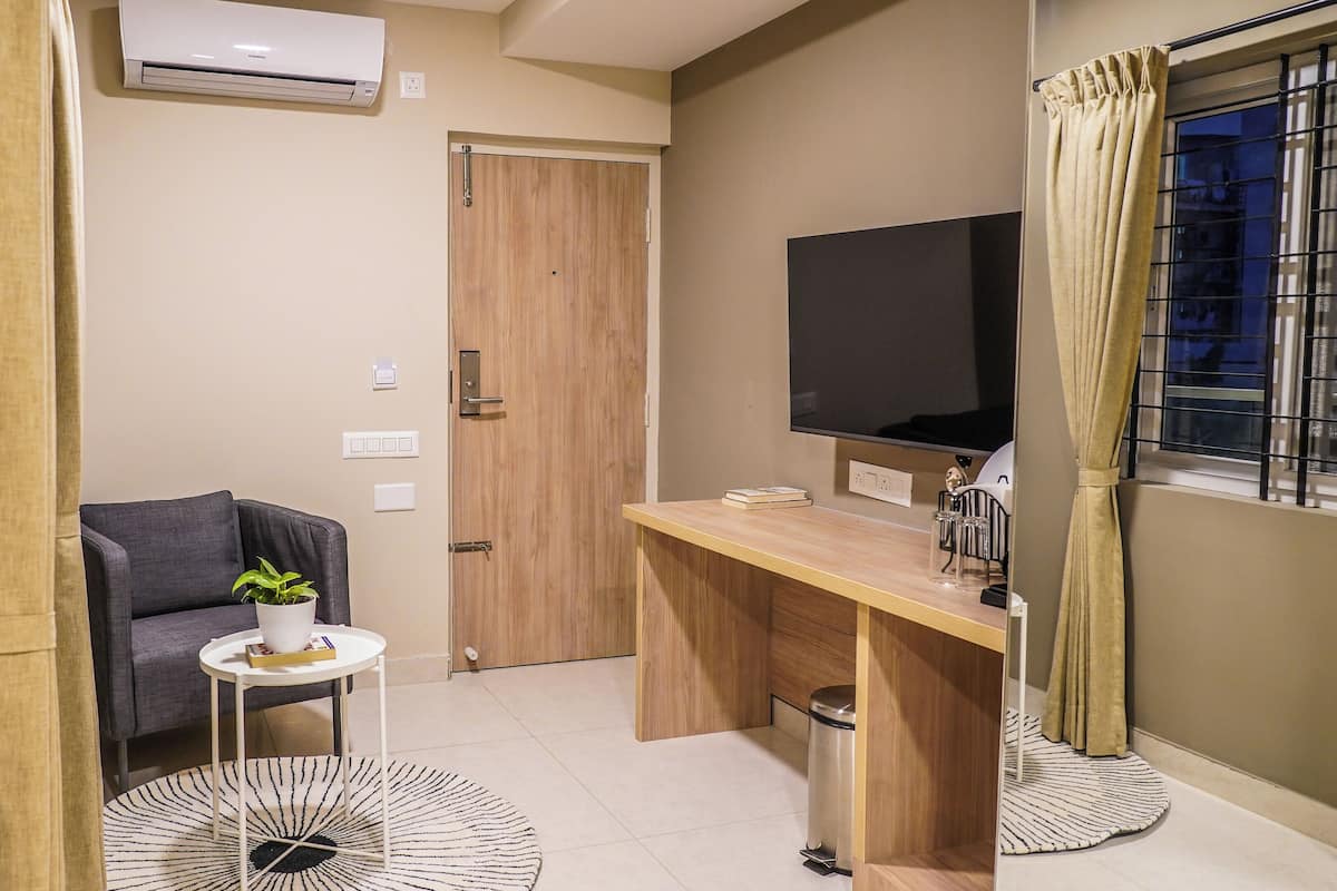 Un espacio habitable pequeño con sofá, una mesita con planta, un escritorio de madera, un televisor y una ventana con cortinas.