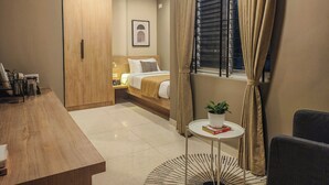 A modern bedroom with a wooden wardrobe, a bed with white bedding, a bedside table with a lamp, a window with curtains, and a small round table with a plant and books.