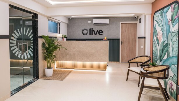 A reception area with a 'live' sign, a reception desk, a potted plant, a bench, and two wicker chairs.