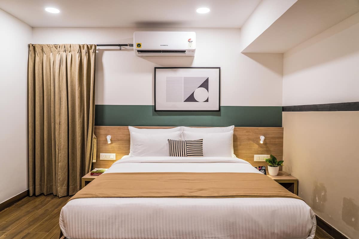 Habitación de hotel con cama, mesita de noche, aire acondicionado instalado en la pared, un cuadro enmarco y cortina.
