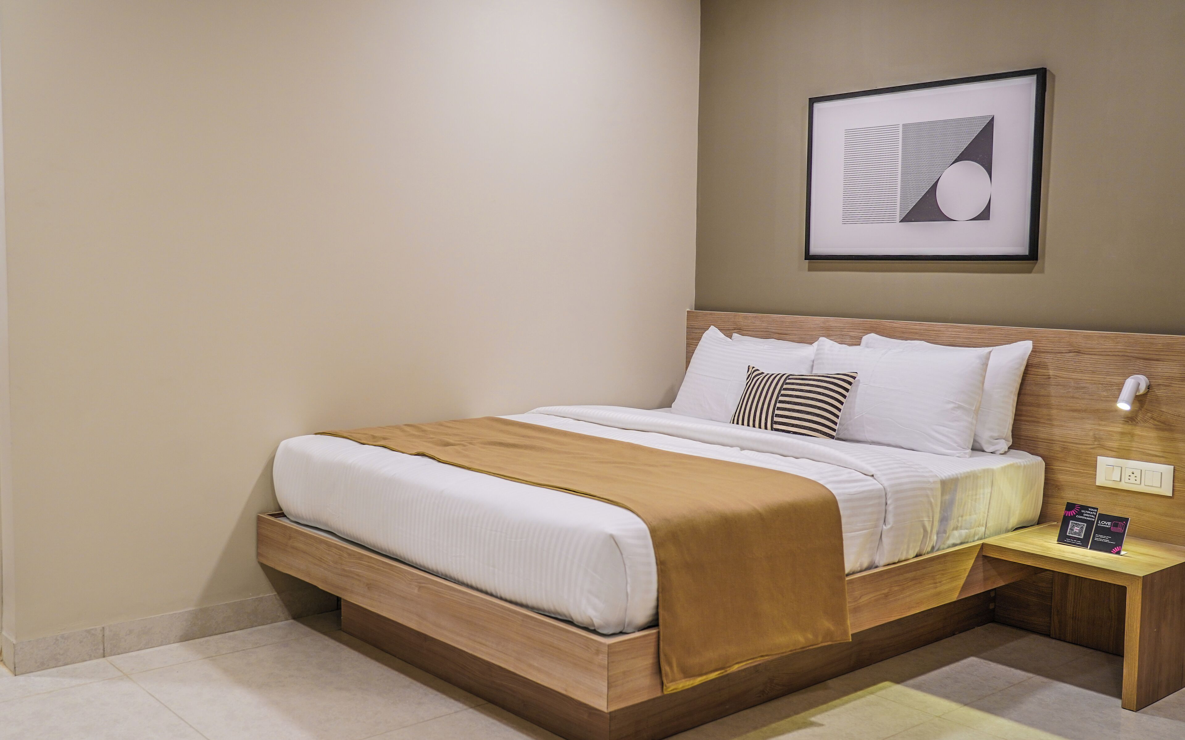 A neatly made bed with a wooden headboard, a bedside table with a lamp, and a framed abstract artwork on the wall.