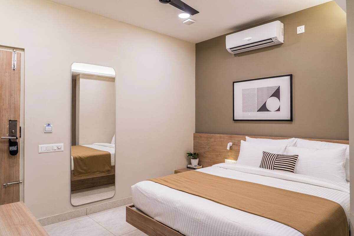 Una habitación de hotel moderna con una cama grande, una mesita de noche con lámpara, aire acondicionado de pared y un cuadro enmarcado sobre la cama.