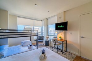A hotel room with bunk beds, a desk with a chair, a television, and a view of the city.