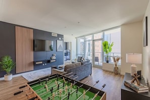 A modern living room with a foosball table, a sofa, a TV mounted on the wall, and large windows providing a view of the city.