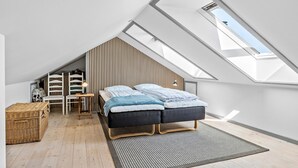 A modern loft bedroom with a sloped ceiling, a bed with a dark headboard, a wooden chair, a small table with a lamp, and a large window providing natural light. - 2 bedroom amazing home in Svaneke (Svaneke)
