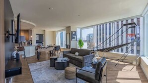 A modern living room with a sofa, coffee table, and ottoman, a dining area with chairs, and a kitchen in the background. - DT PHX Stay | Pool, Gym, Balcony, W/D + Parking (Phoenix)