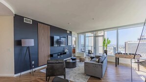 A modern living room with a large TV, comfortable seating, a foosball table, and a city view through large windows. - Stadium District Apt Pool,Gym,Balcony,Parking,WD (Phoenix)