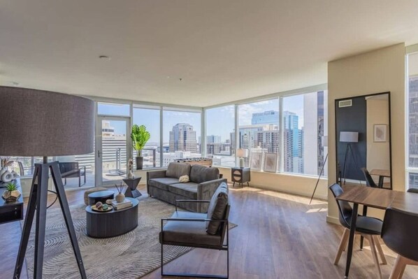A spacious living room with a large window, a grey sofa, a coffee table, and a dining area with a table and chairs. - Stadium District Apt Pool,Gym,Balcony,Parking,WD (Phoenix)
