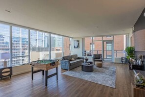 A spacious living area with a foosball table, a grey sofa, and a coffee table. Large windows offer a view of the cityscape. - Modern ASU Retreat | Pool | Gym | Balcony | W/D (Phoenix)