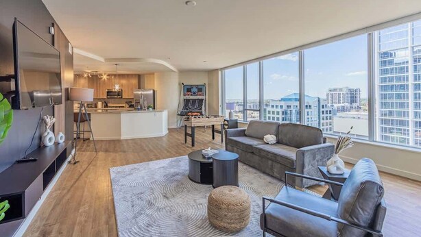 Una sala de estar moderna con sofá, mesa de centro y televisor. Se ve una cocina al fondo con un comedor.