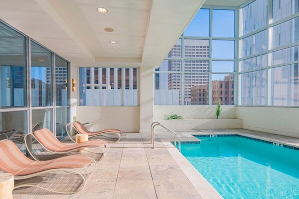 A pool area with lounge chairs, a pool, and large windows offering a city view.
