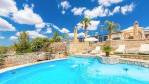 A swimming pool with stone walls and lounge chairs, surrounded by palm trees and a clear blue sky.