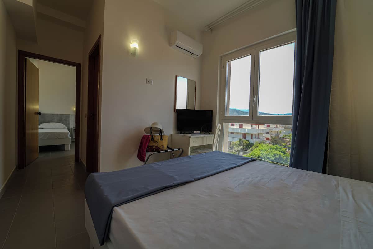 A hotel room with a bed, a desk with a television, a window with a view, and an air conditioning unit.