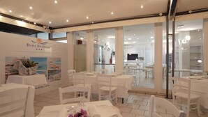 A hotel restaurant with white tables and chairs, a TV in the background, and a sign for Hotel Rivabella. - Hotel Rivabella (Davoli)