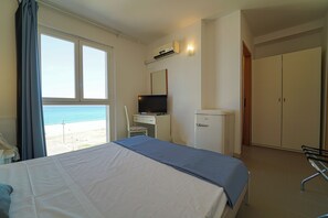 A hotel room with a bed, a desk with a computer, a television, a wardrobe, and a view of the beach.