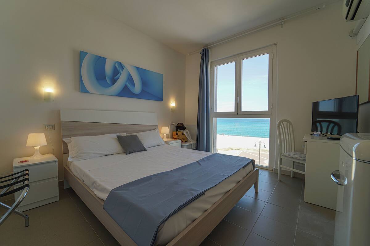 A hotel room with a bed, a desk, a chair, a TV, and a window with a view of the beach.