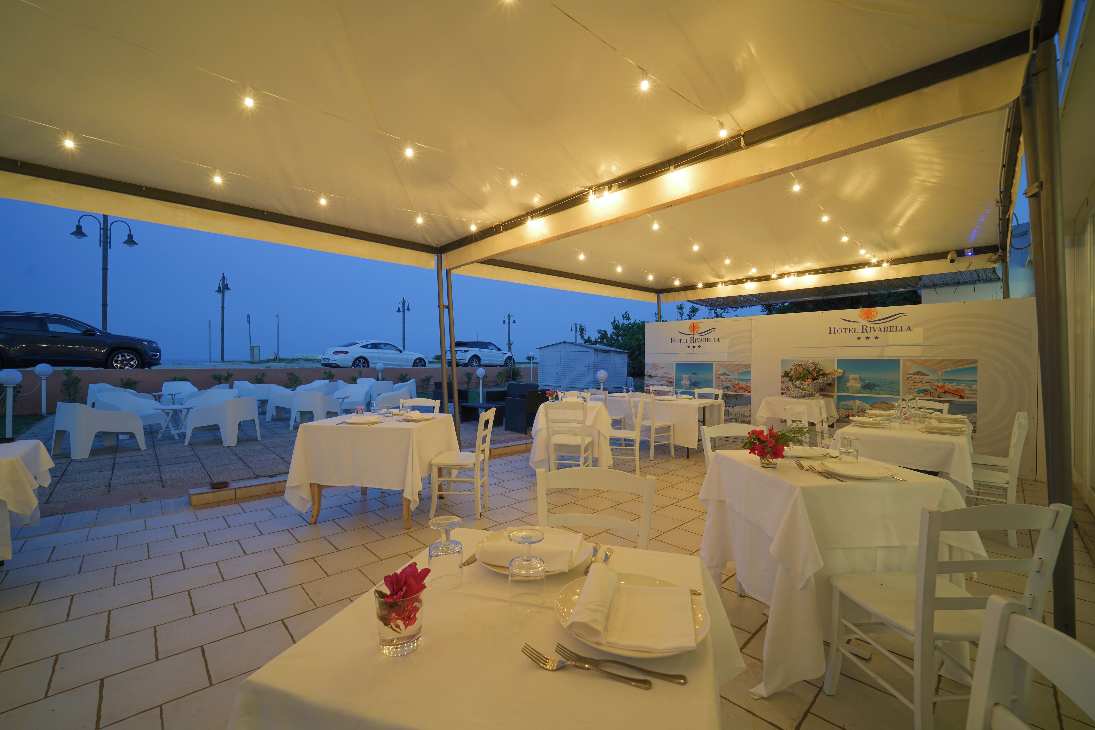 An outdoor dining area with white tables and chairs, a covered section with string lights, and a view of parked cars and a building in the background.