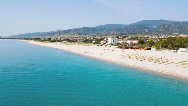 A coastal beach with clear turquoise waters, a sandy shore, and a backdrop of hills and buildings. - Hotel Rivabella (Davoli)