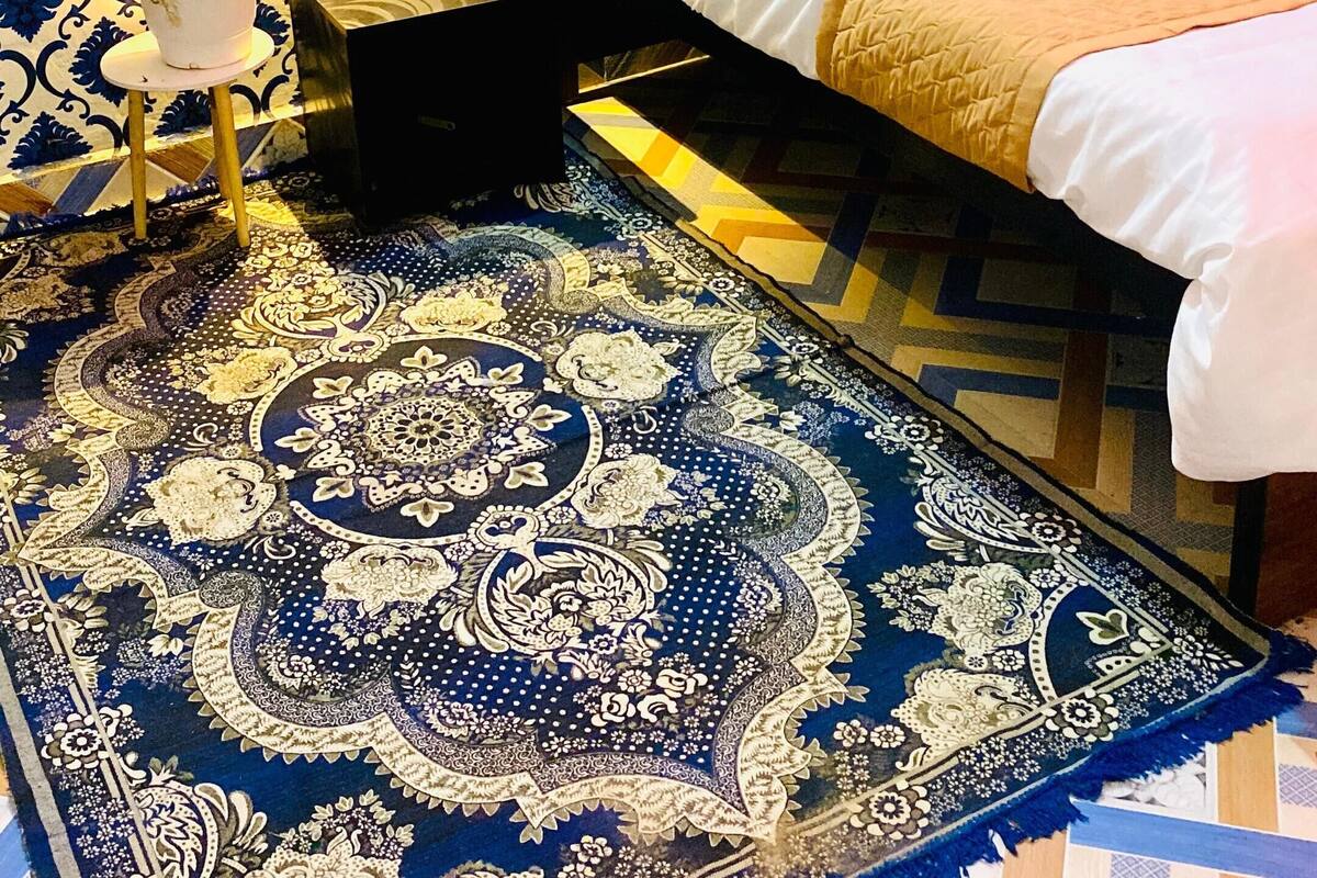 Une chambre à coucher avec un lit, des oreillers, une table de chevet, une lampe, une plante en pot et un tapis à motifs.