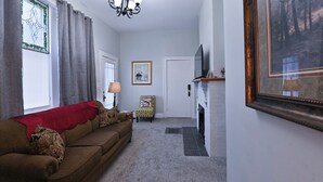 A living room with a brown sofa, a small table, a lamp, a framed picture, a fireplace, and a TV on a stand. - Dual patios, Free Laundry Service, College steps, 2BR 1 BA (Atchison)