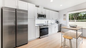 A modern kitchen with white cabinets, stainless steel appliances, a wooden table, and a window with a view of trees.