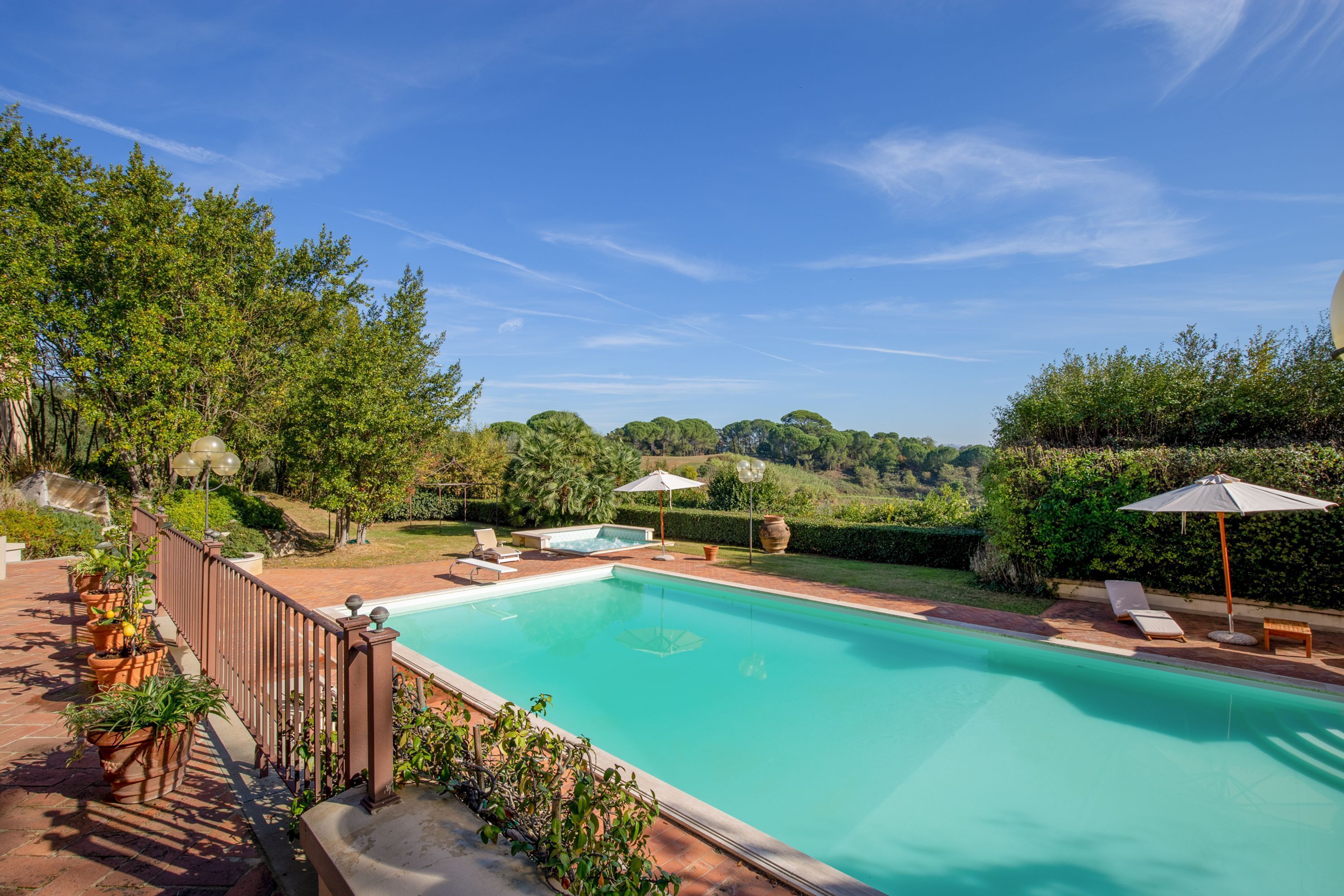 A swimming pool with lounge chairs and umbrellas, surrounded by greenery and a clear sky.