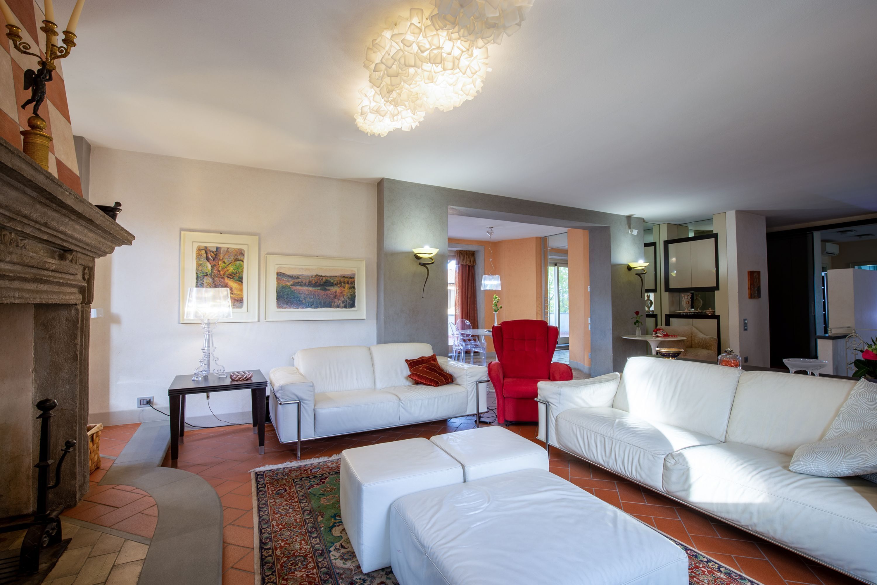 Een woonkamer met een open haard, witte banken, een rode fauteuil en een kroonluchter.
