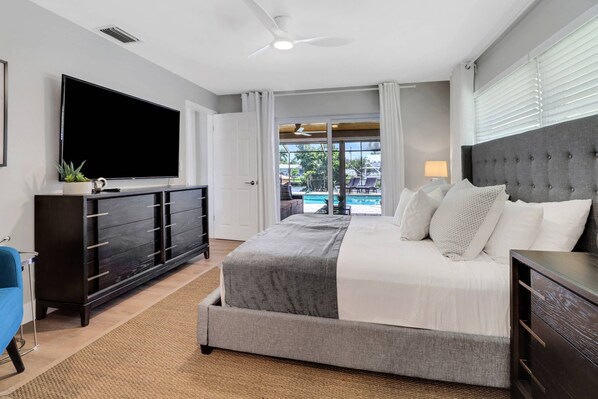 Une chambre d’hôtel moderne dotée d’un grand lit, d’une télévision, d’une commode en bois foncé et offrant une vue sur une piscine et un espace extérieur.