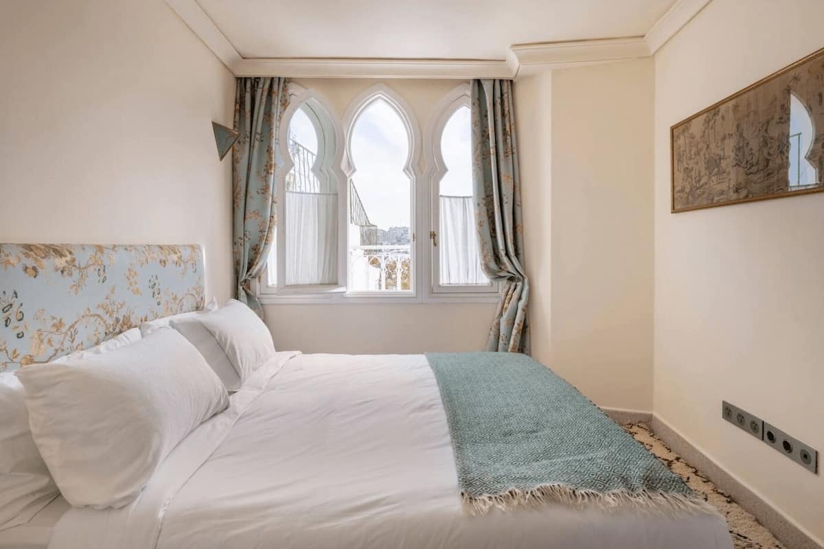 A bedroom with a bed, two windows with curtains, a wall-mounted artwork, and a radiator.