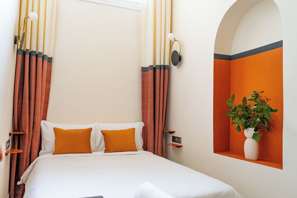 A neatly made bed with white and orange bedding, a bedside table with a lamp, and a wall-mounted shelf with a plant.