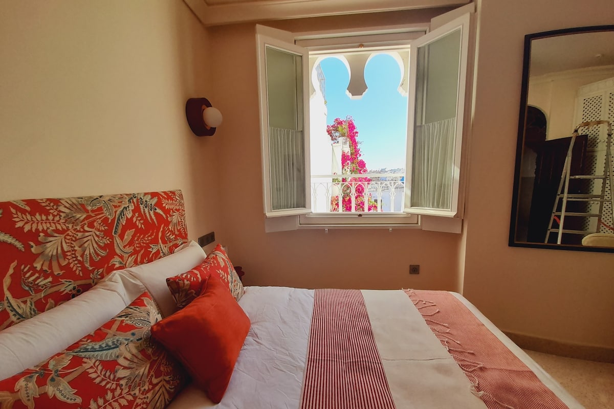 A bedroom with a bed, a window with a view of a balcony, and a mirror on the wall.
