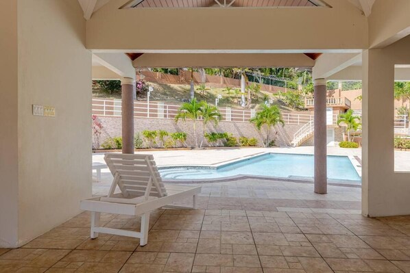A covered pool area with a white lounge chair, tiled flooring, and a view of a landscaped garden with palm trees and a building in the background.