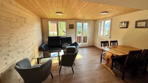 Un amplio salón con techo de madera, un sofá negro, una mesa de centro de vidrio y una mesa de comedor de madera con sillas.