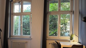 A bedroom with a bed, a desk, and a chair. There are two windows with views of trees. - Blue Doors Hostel Altstadt (Rostock)