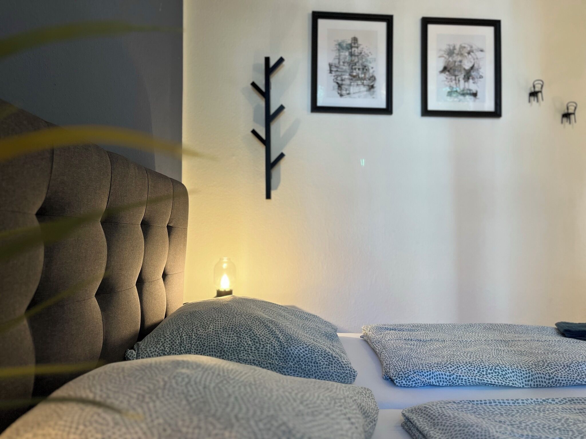 Un dormitorio con cabecera capitonada, dos almohadas, una mesita de noche con vela y dos obras de arte enmarcadas en la pared.