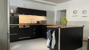 A modern kitchen with black cabinets, stainless steel appliances, and a wooden countertop. There are two clocks on the wall and a potted plant on the island. - Blue Doors Hostel Altstadt (Rostock)
