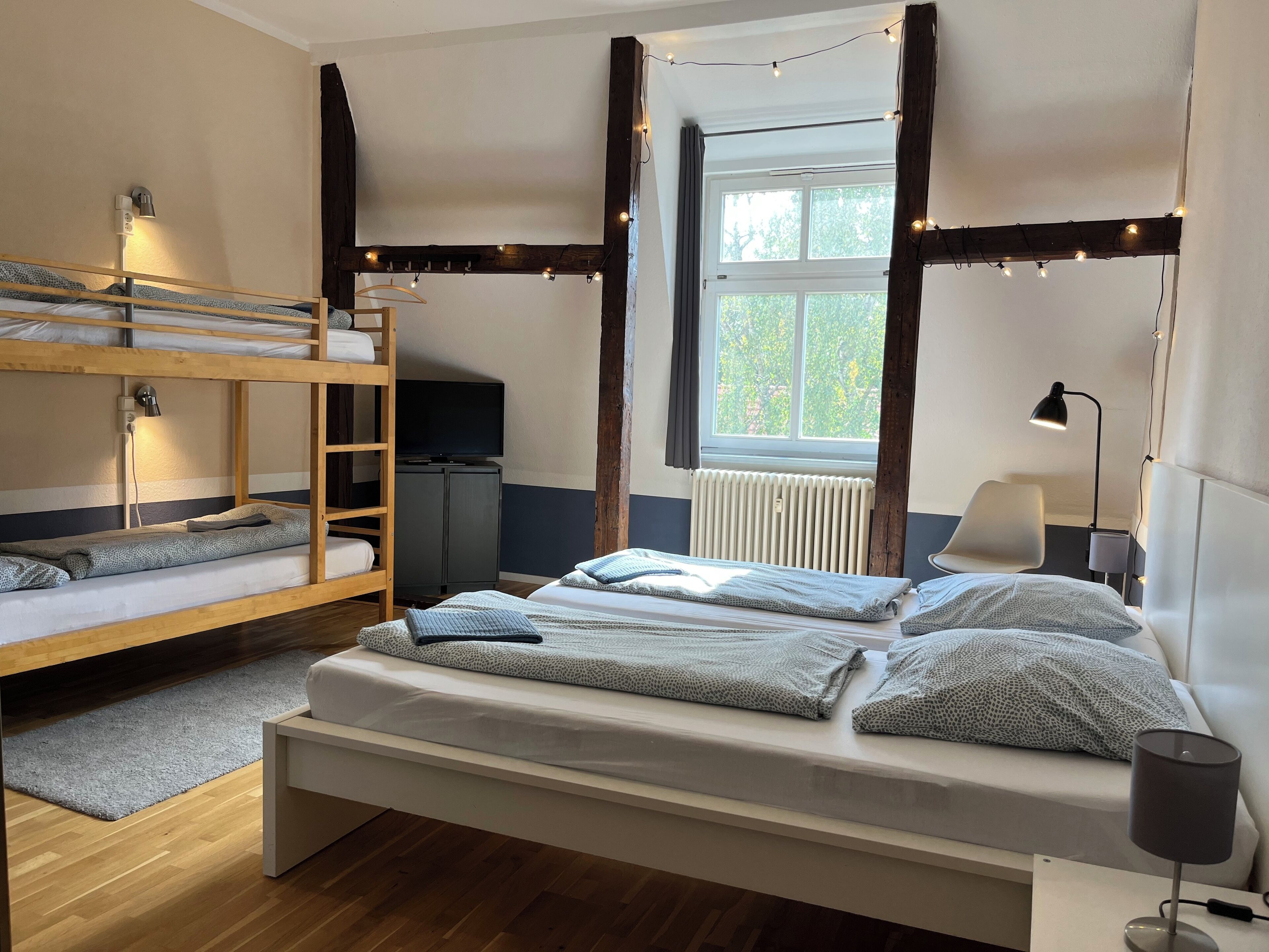 A dormitory room with bunk beds, a television, a window with curtains, and string lights.