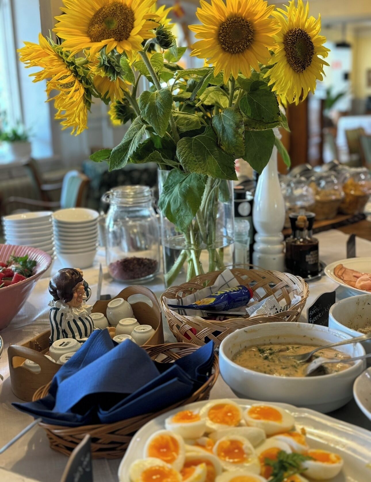 Una mesa dispuesta con diferentes platos, incluyendo una cesta de huevos, un tazón de sopa y un florero con girasoles.