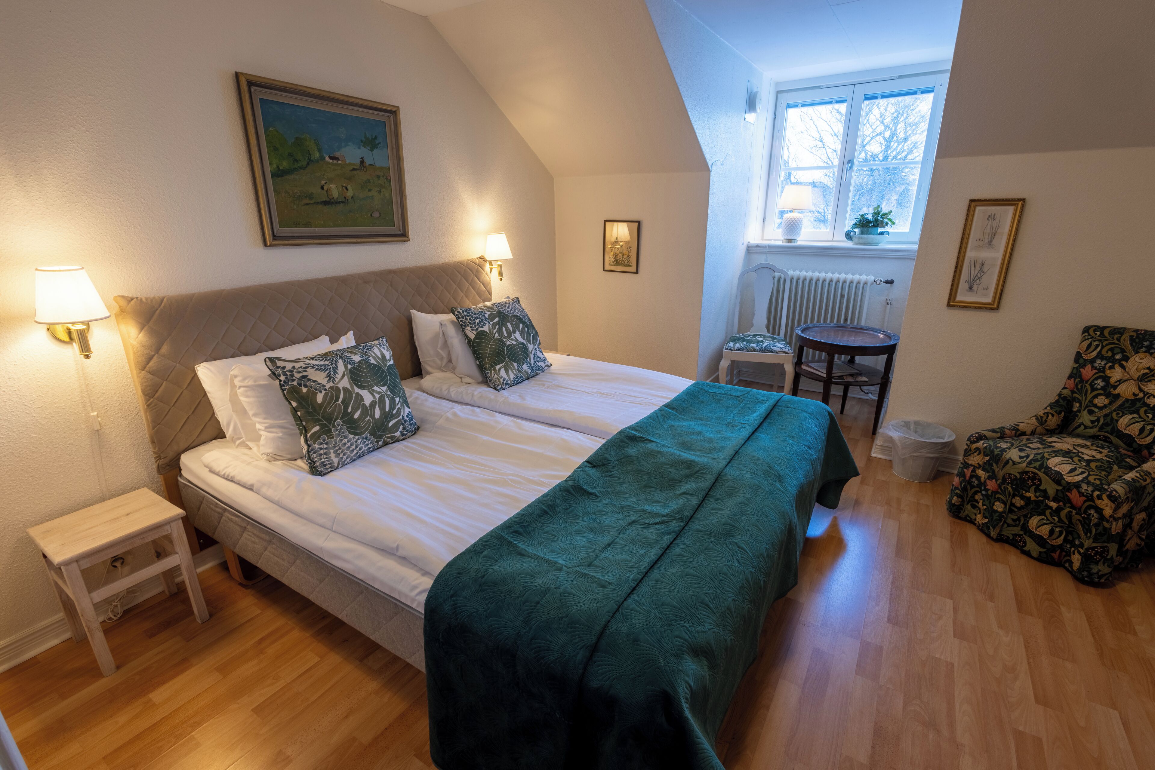 A bedroom with a bed, two bedside lamps, a small table, a chair, and a window with a view of the outside.