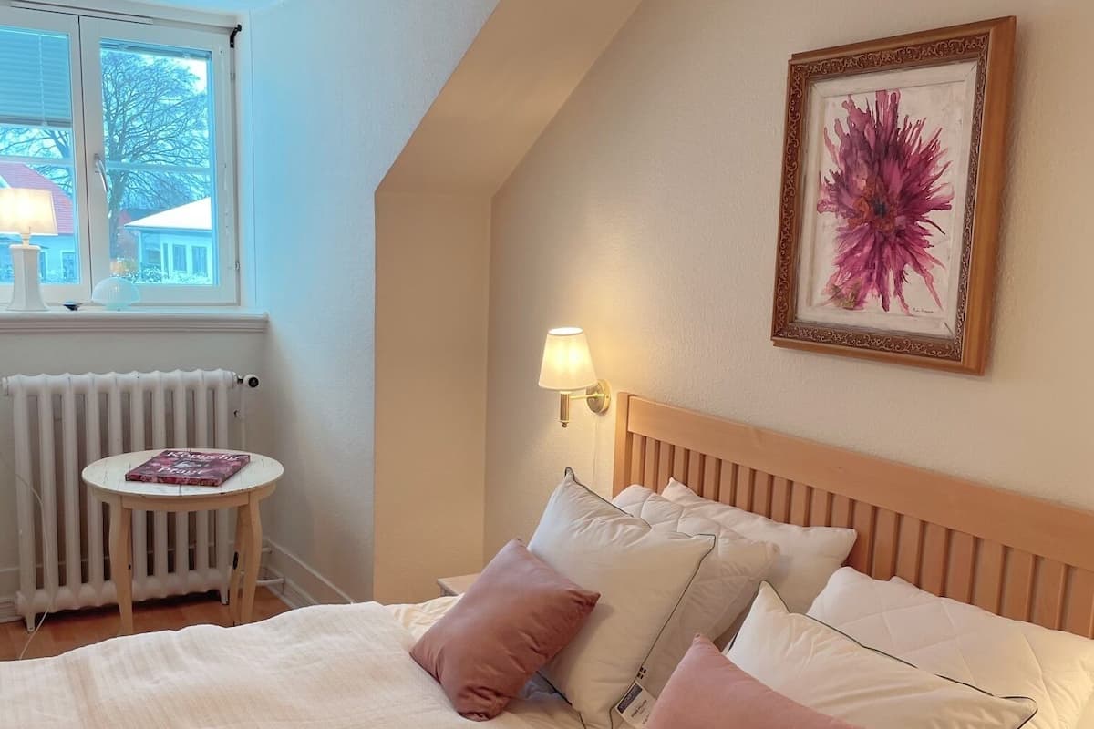 Un dormitorio con cama, almohadas, una mesita de noche, lámpara, radiador y ventana con vistas a edificios.