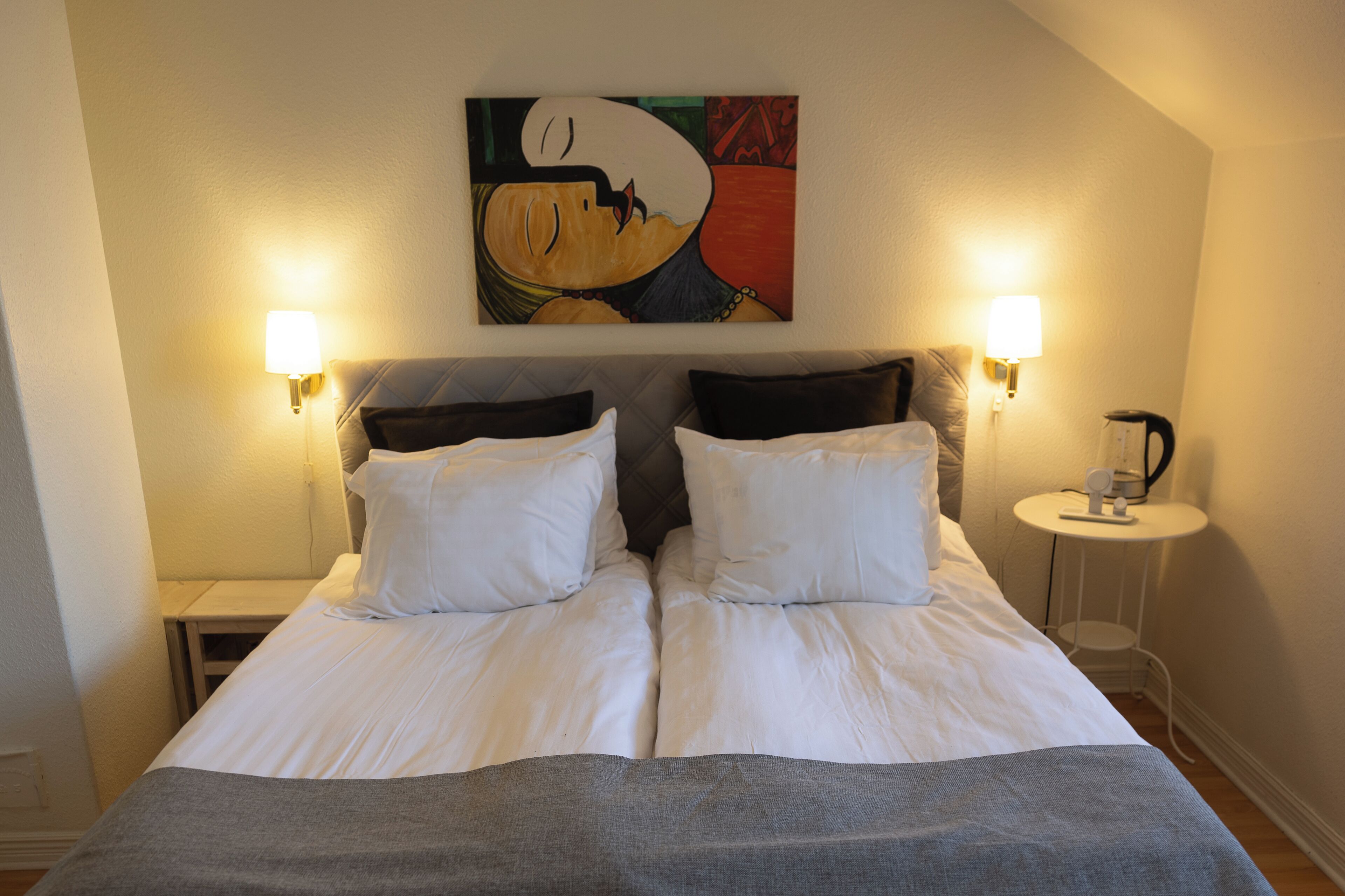 A double bed with white linens, two bedside lamps, and a painting above it.