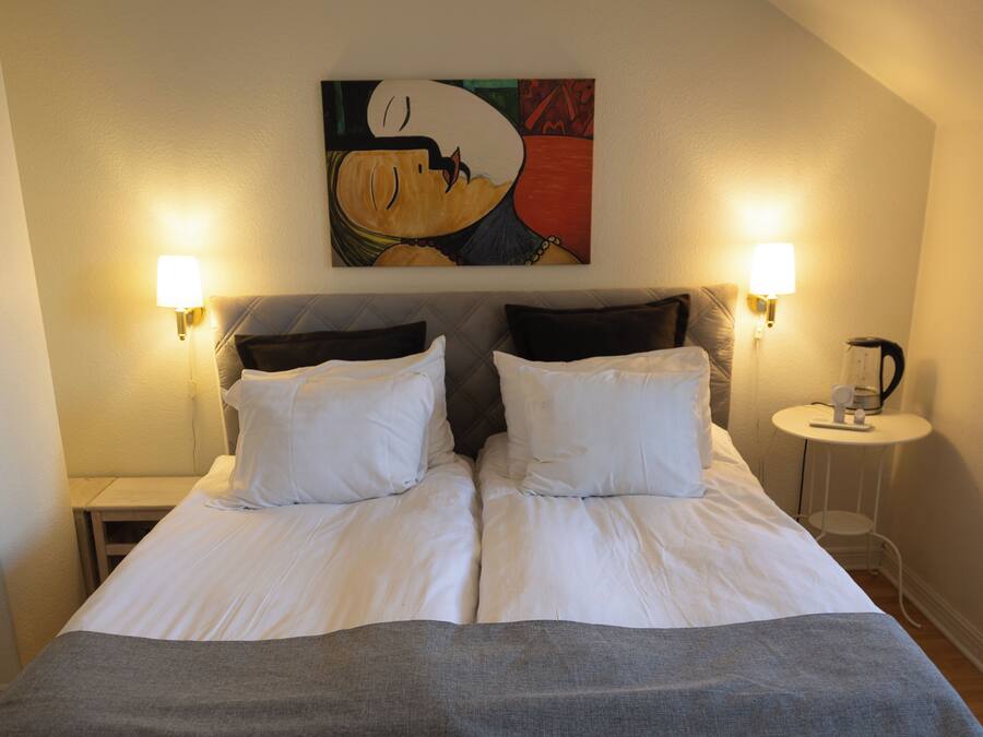 A double bed with white linens, two bedside lamps, and a painting above it.