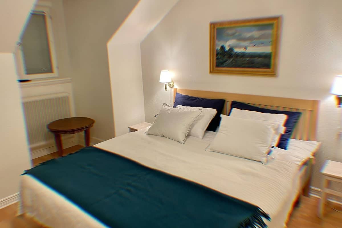 Una cama bien hecha con sábanas blancas y una manta color turquesa, una mesita de noche con lámpara, y un cuadro en la pared.