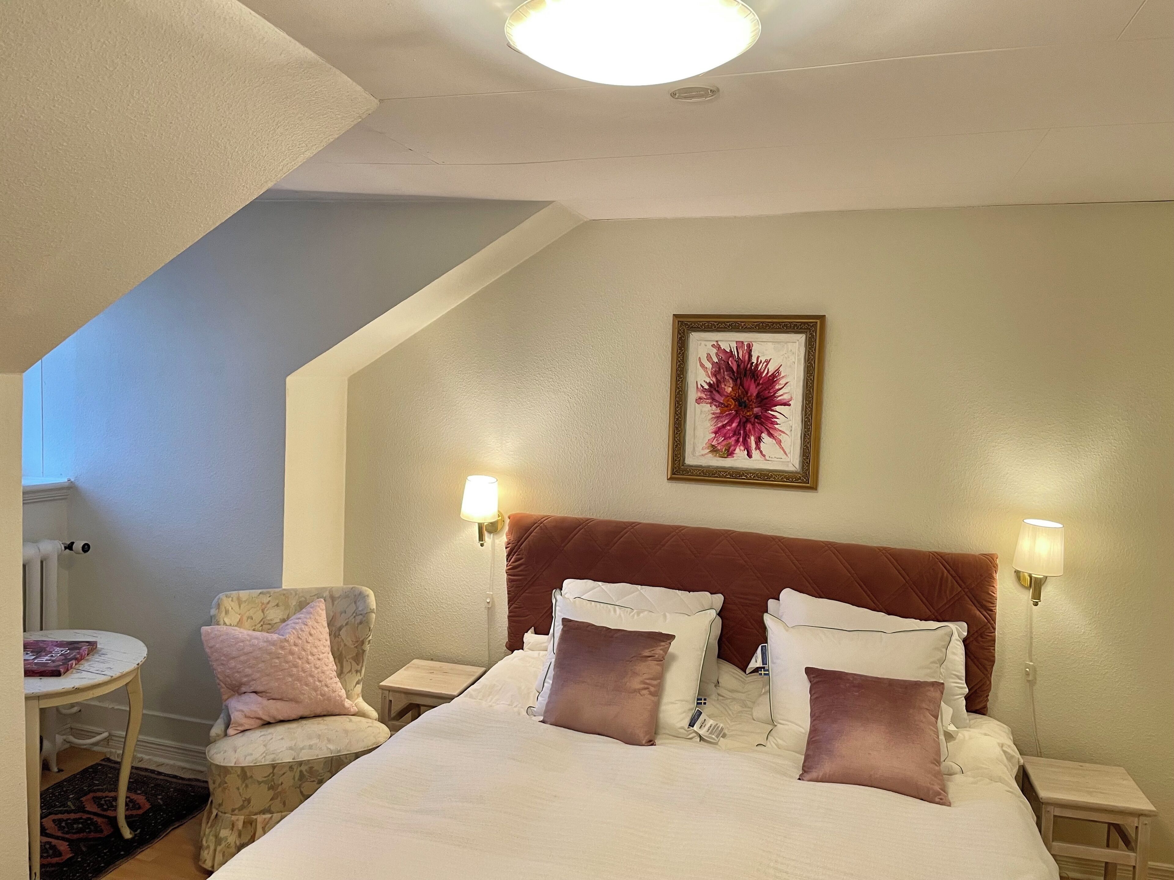A bedroom with a bed, two bedside lamps, a chair, a small table, and a framed artwork on the wall.