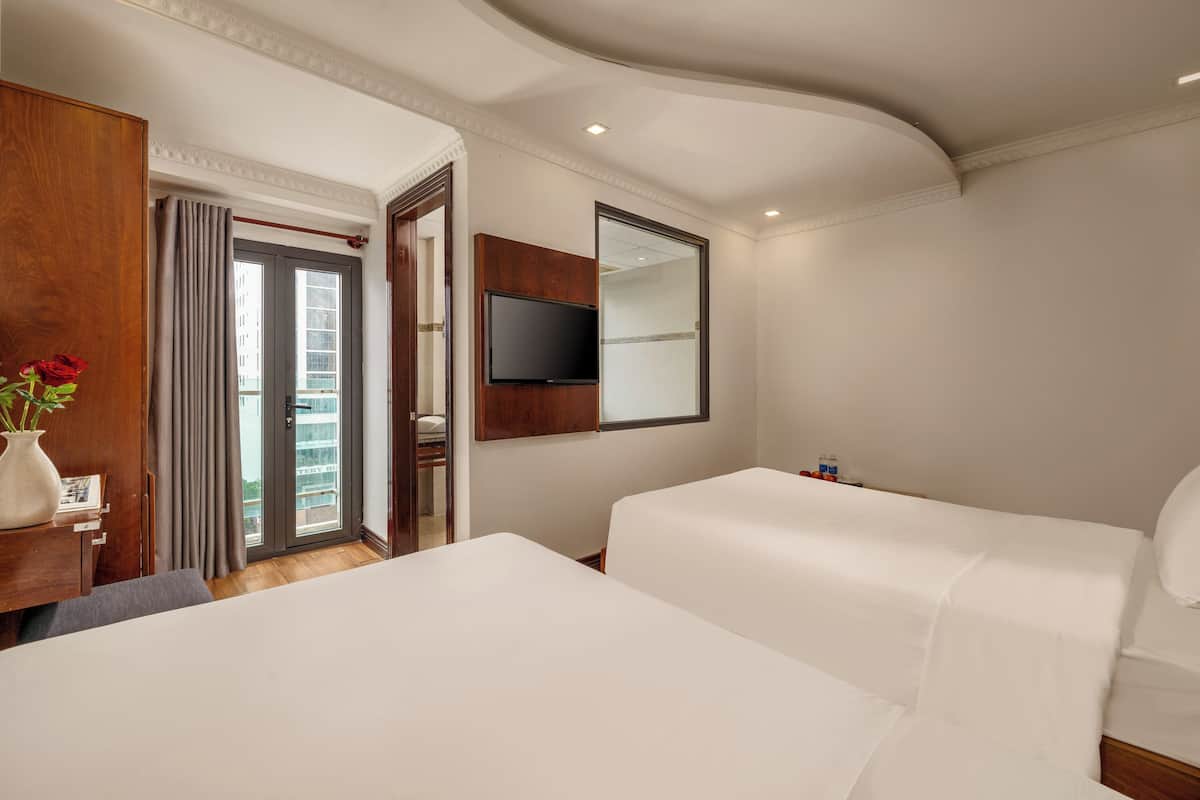 Une chambre d’hôtel avec deux lits, un téléviseur fixé au mur, un bureau en bois agrémenté d’un vase de fleurs et une porte-fenêtre coulissante donnant sur un balcon.