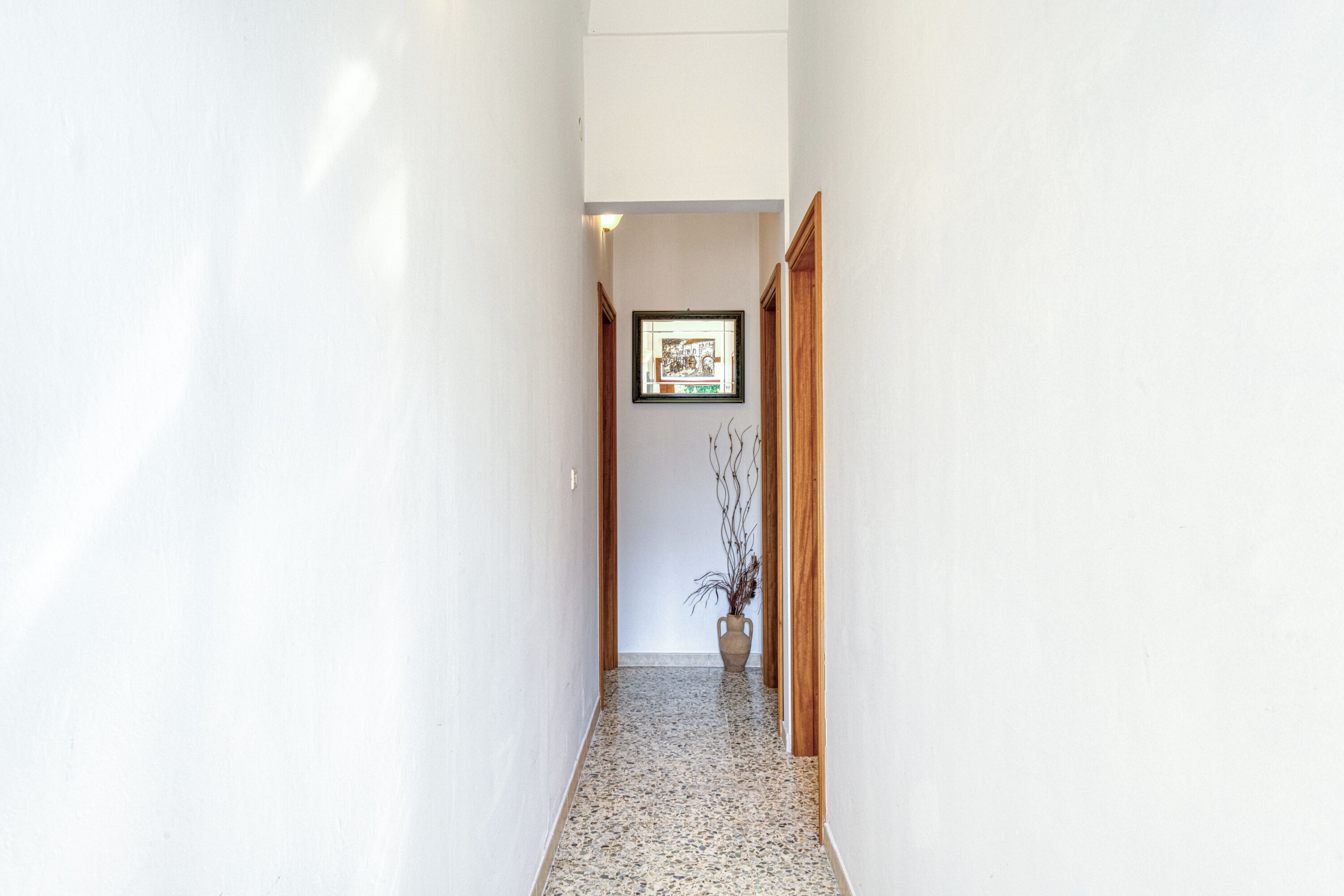 Un corridoio stretto con pareti bianche e pavimento piastrellato, che conduce a una porta in fondo.