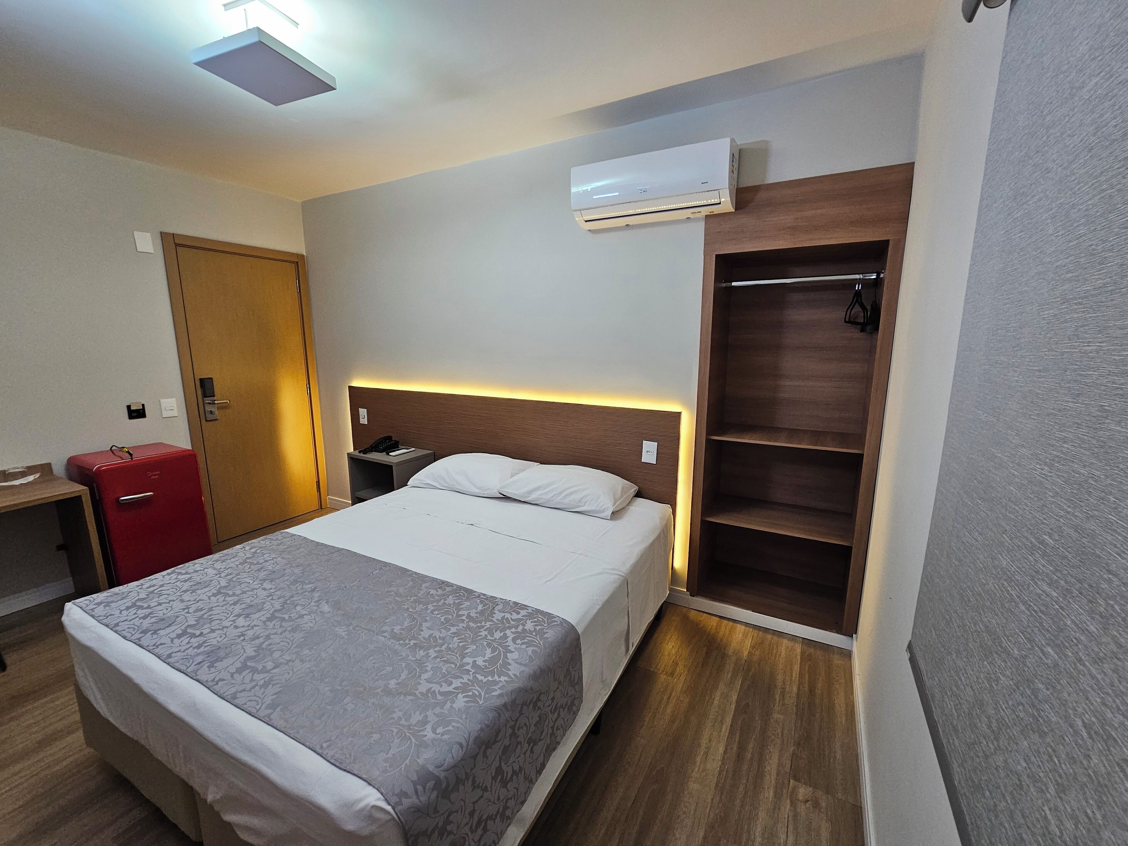 A hotel room with a bed, bedside table, a red suitcase, an air conditioning unit, and a wooden wardrobe.