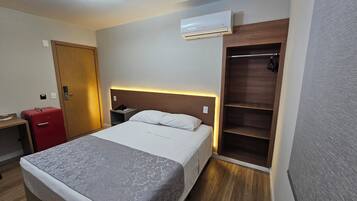 A hotel room with a bed, bedside table, a red suitcase, an air conditioning unit, and a wooden wardrobe.