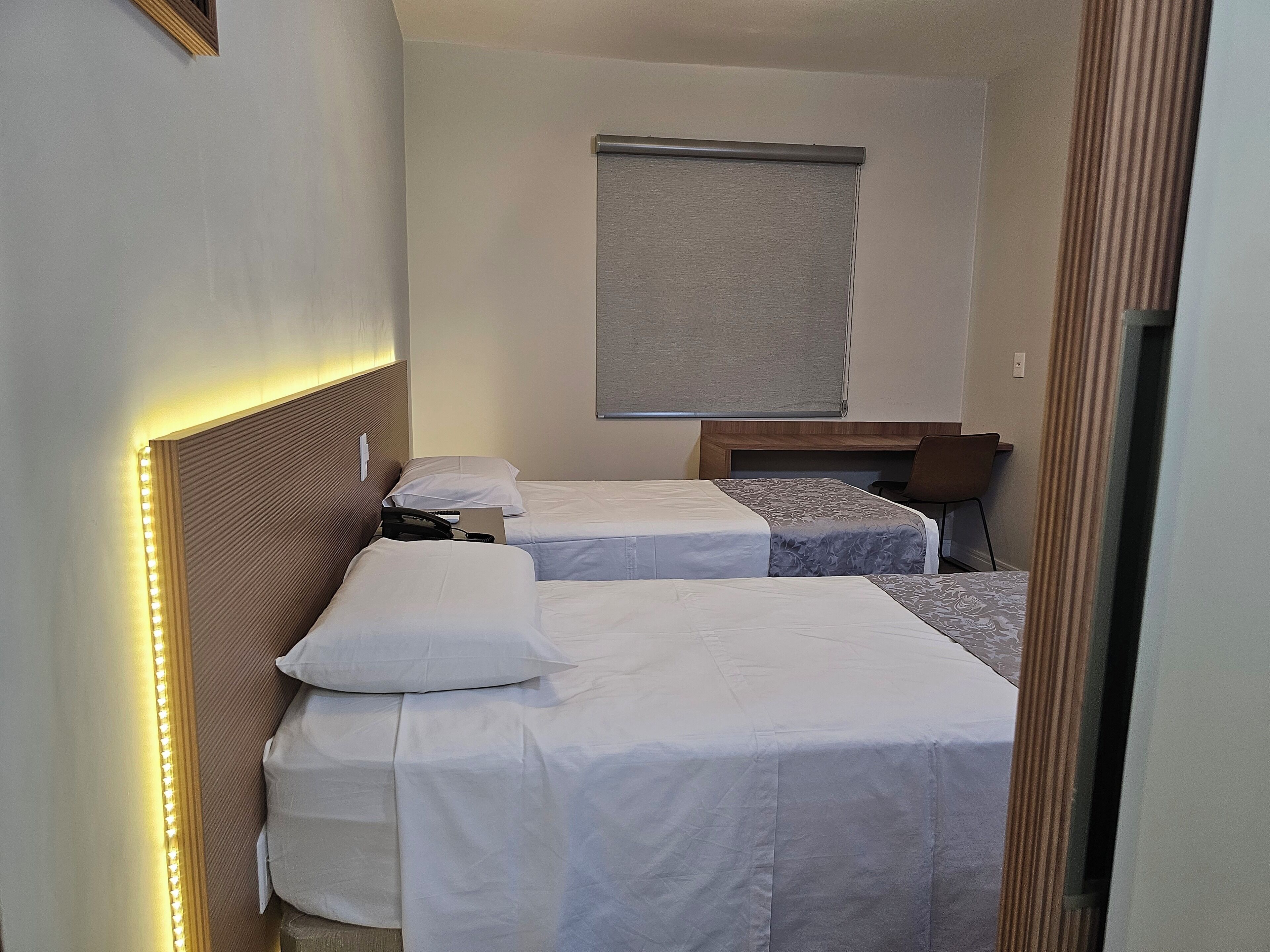 Habitación de hotel con dos camas individuales, una mesita de noche con lámpara, una silla y una ventana con persiana.